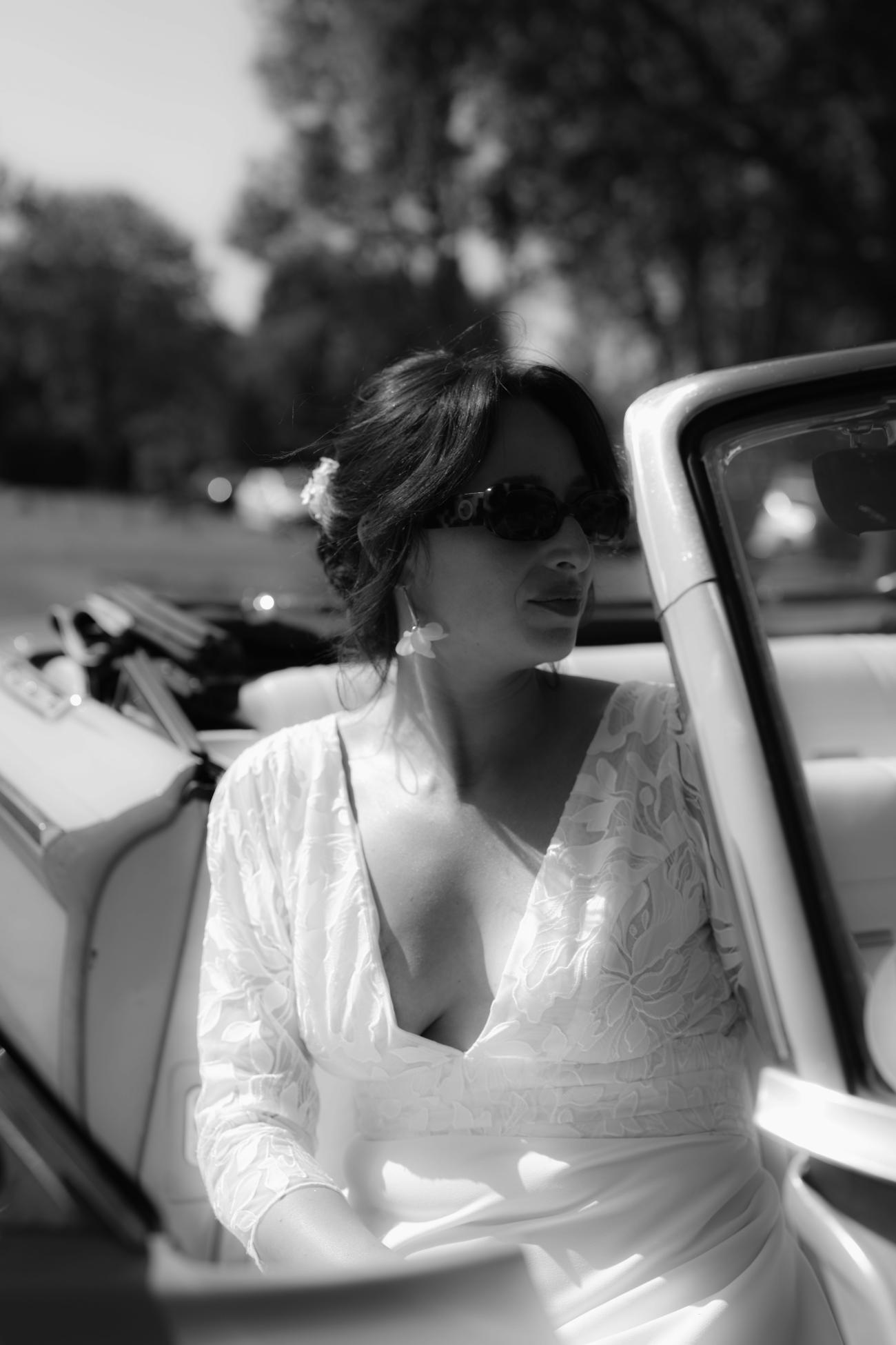 Mariée élégante dans une voiture ancienne Mercury au Domaine de la Métairie à Ossun : photo de mariage en noir et blanc au rendu vaporeux.